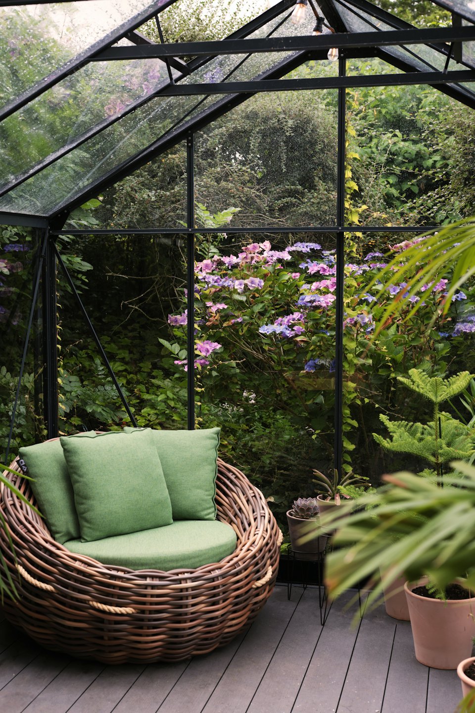 Wicker chair with green cushions inside a modern glass greenhouse.